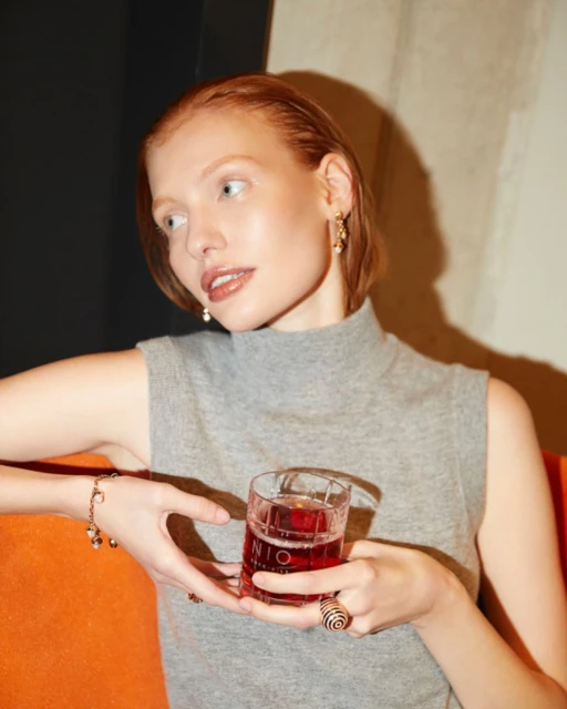 Young adult woman with red hair holds a Nio cocktail in a glass. She wears jewelry and a grey top, sitting on an orange sofa.