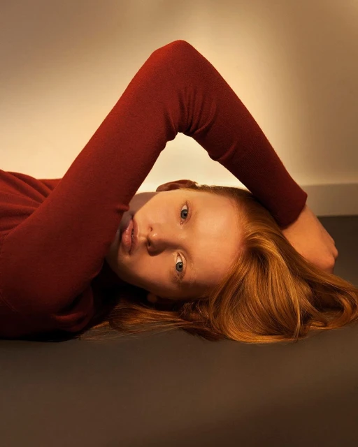 Portrait of a red-haired female model with fair skin lying on her back in a rust-colored sweater, posing against a neutral backdrop.