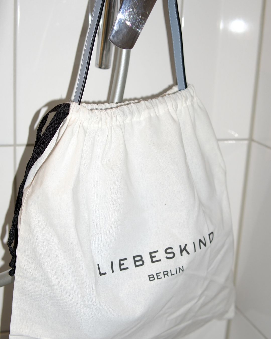 White Liebeskind Berlin drawstring bag hanging on a silver metal bar against a white tiled wall in an indoor setting.