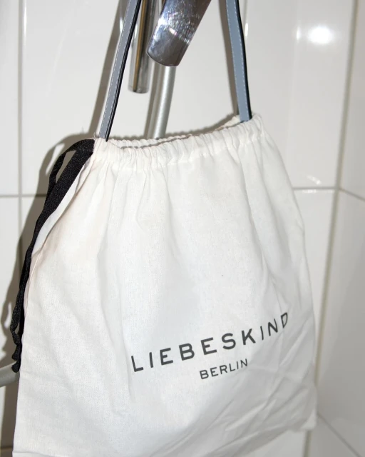 White Liebeskind Berlin drawstring bag hanging on a silver metal bar against a white tiled wall in an indoor setting.