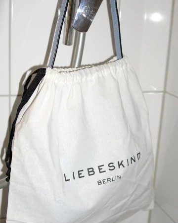 White Liebeskind Berlin drawstring bag hanging on a silver metal bar against a white tiled wall in an indoor setting.
