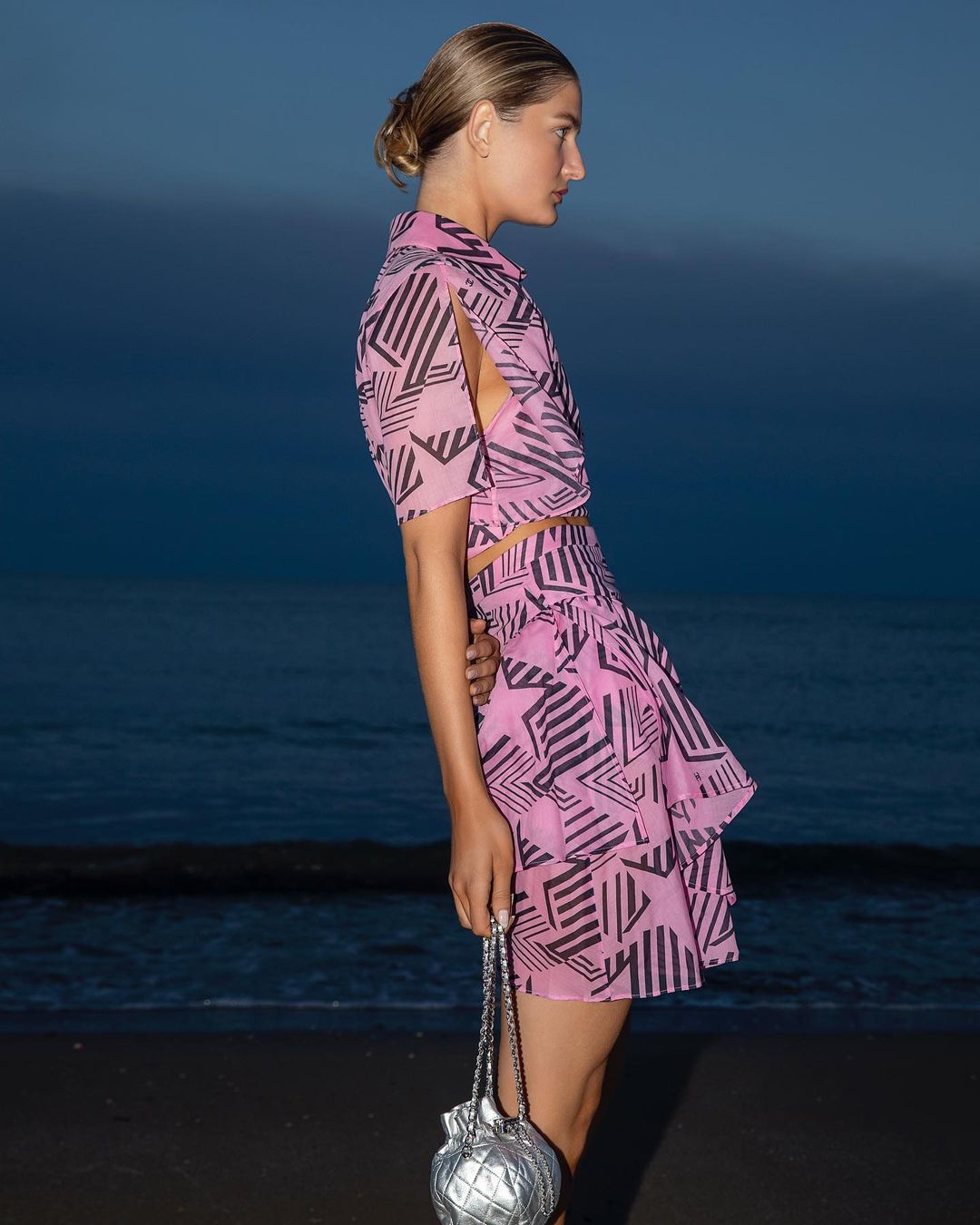 Profile view of a fair-skinned adult woman in a patterned pink two-piece outfit with a silver purse, posing on a beach at dusk.