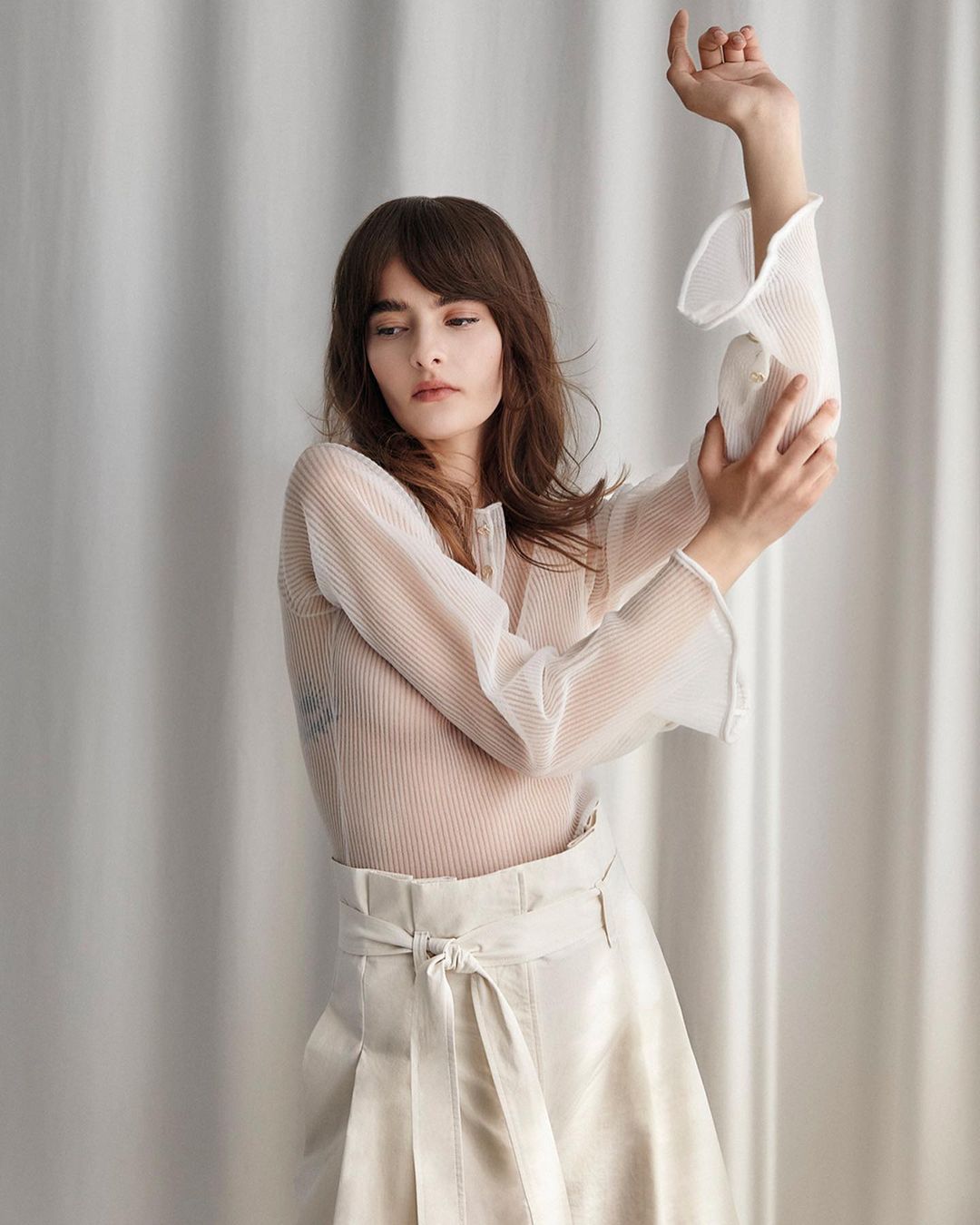 Portrait of a woman in a white sheer blouse and white skirt posing against a white draped fabric background in a photo studio.