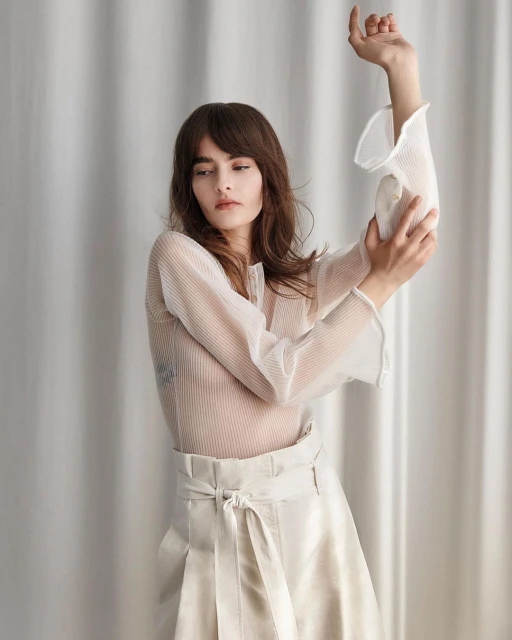 Portrait of a woman in a white sheer blouse and white skirt posing against a white draped fabric background in a photo studio.