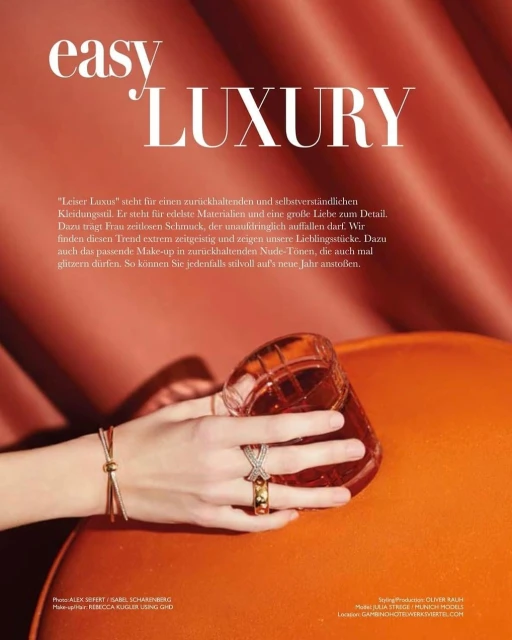Close-up of a hand wearing rings and a bracelet next to a glass on an orange surface, with soft studio lighting and peach-colored background.