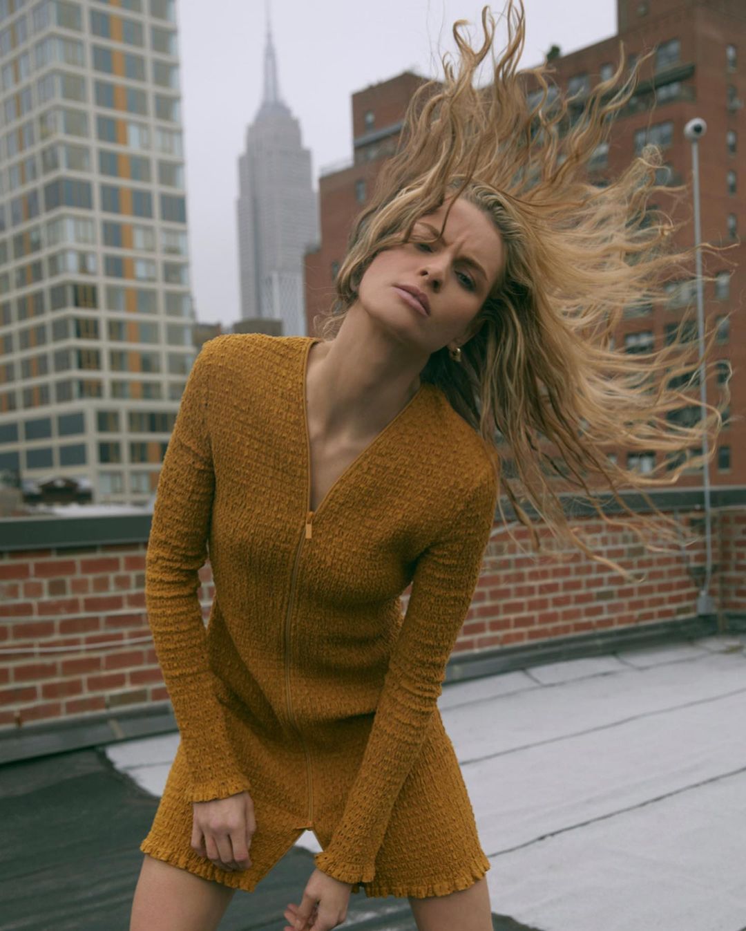 Fashion shot of a blonde adult woman in an orange mini dress with long sleeves, posing on a rooftop in an urban setting with skyscrapers.