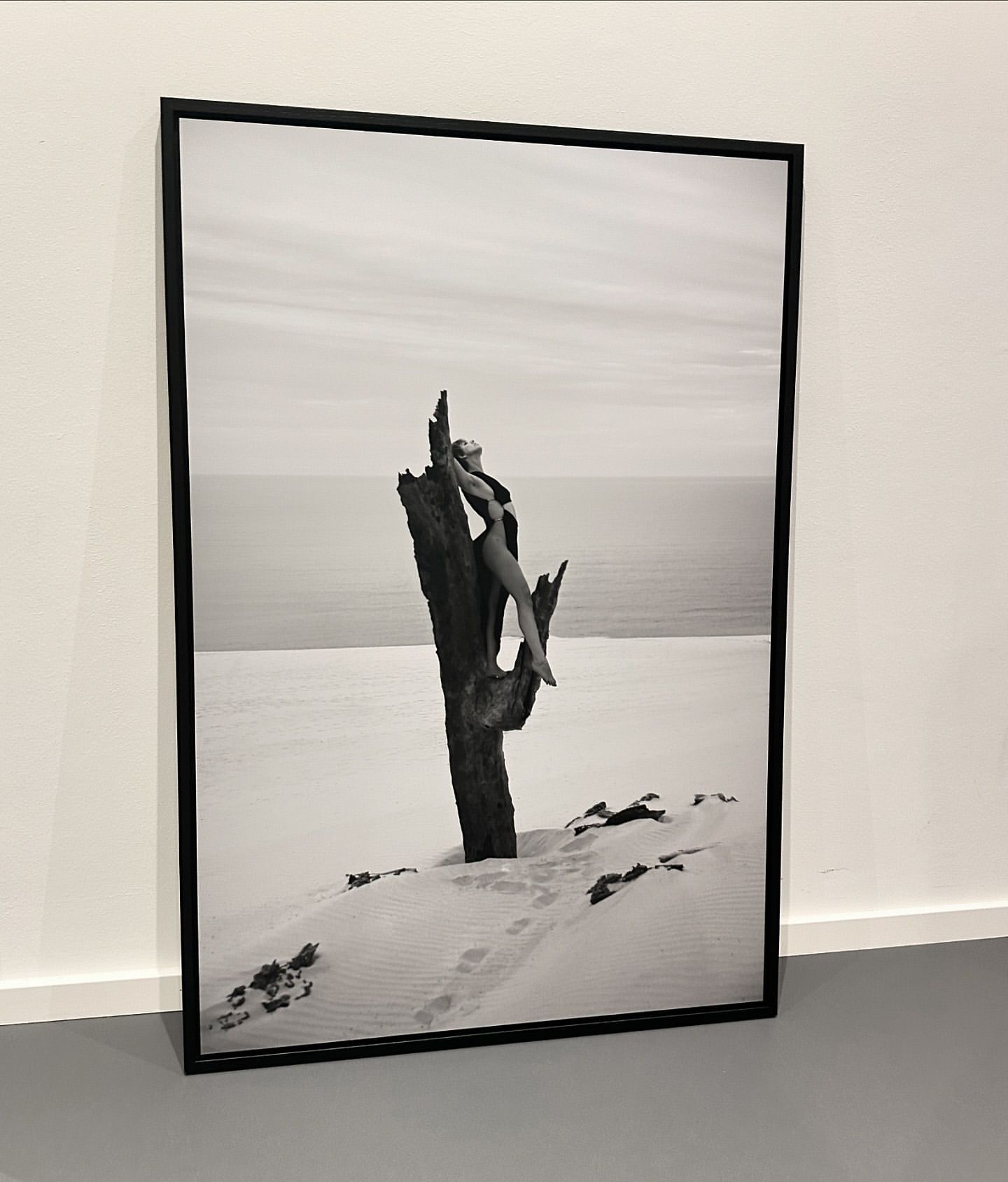 Framed black and white photograph of a woman posing on a weathered tree trunk in a sandy landscape with the ocean behind
