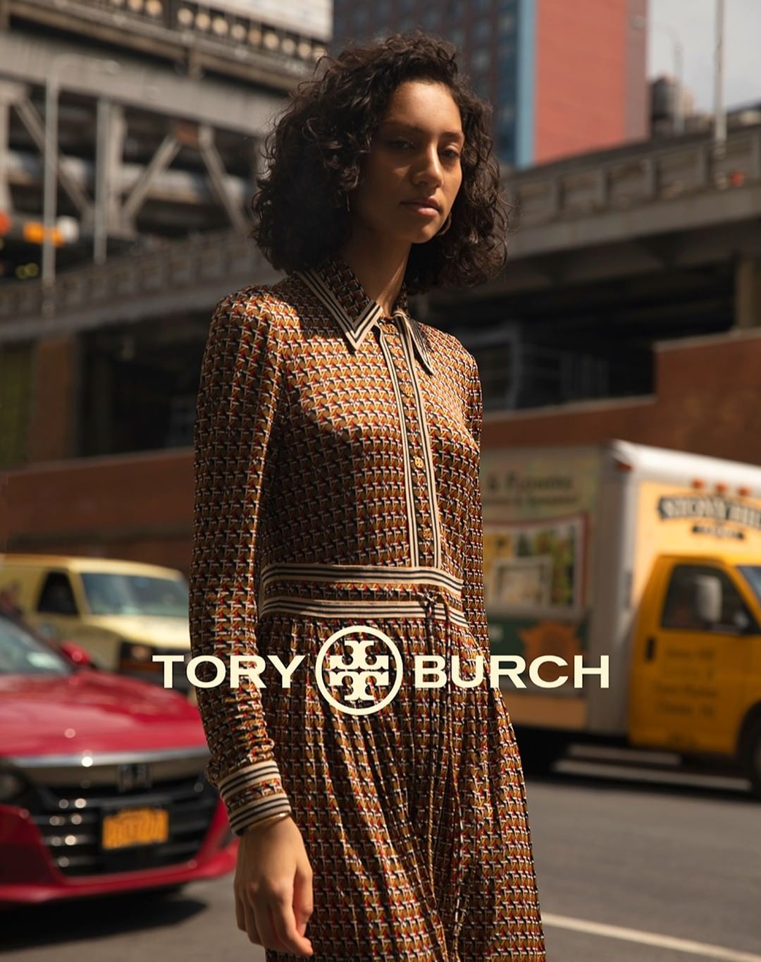An adult female fashion model poses in a Tory Burch patterned dress on a city street with cars and buildings in the background.
