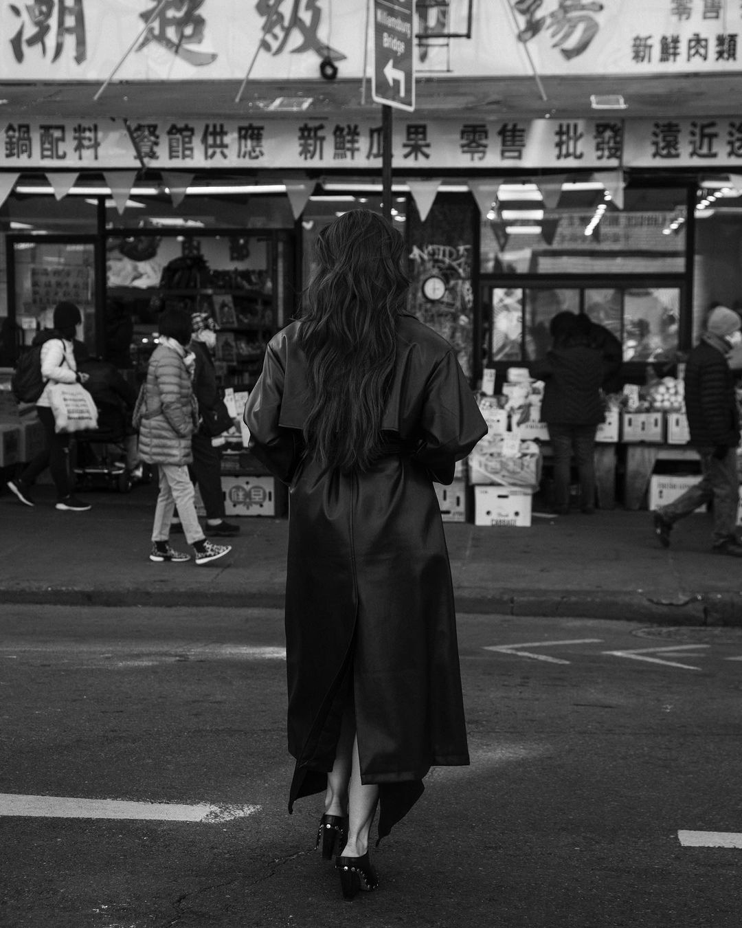 Person in dark coat walking on city street in black and white. Long hair, back view, commercial signage and pedestrians in background.