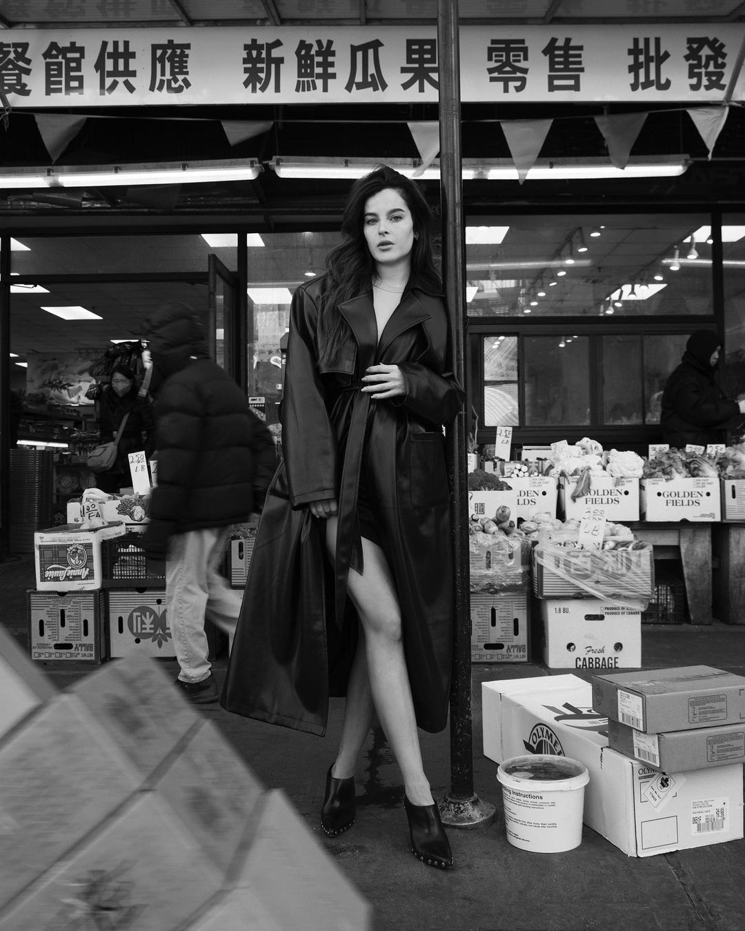 Black and white portrait of a woman in a trench coat standing in front of a storefront with food crates in an outdoor, urban environment.