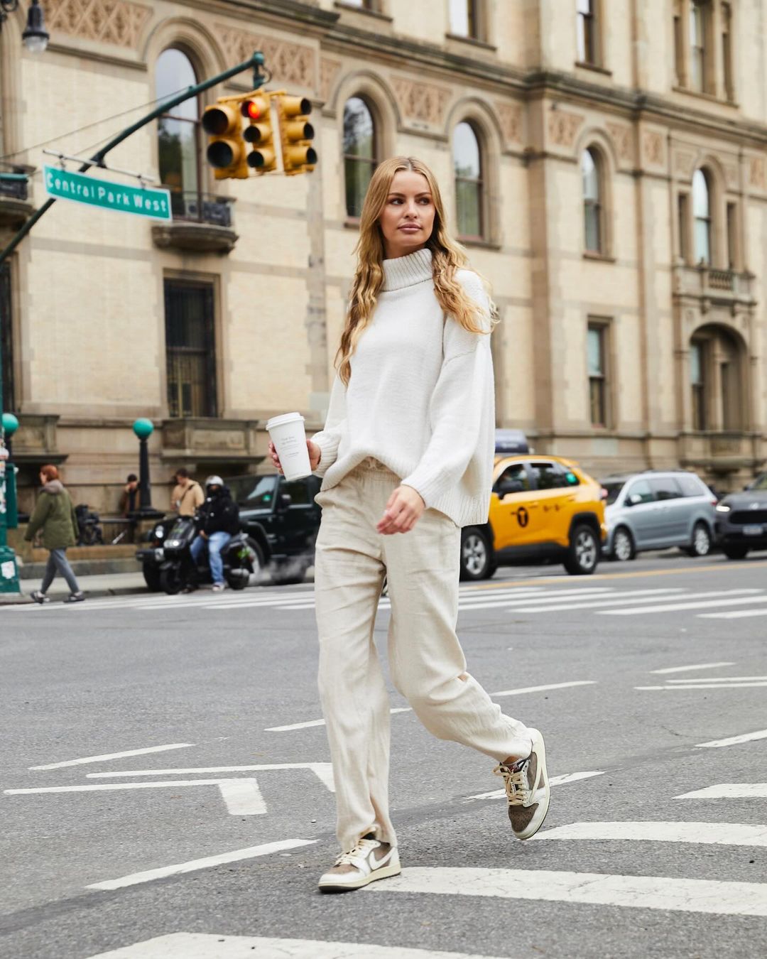 Woman in a white sweater and beige pants walking across a crosswalk in an urban setting with buildings and traffic.