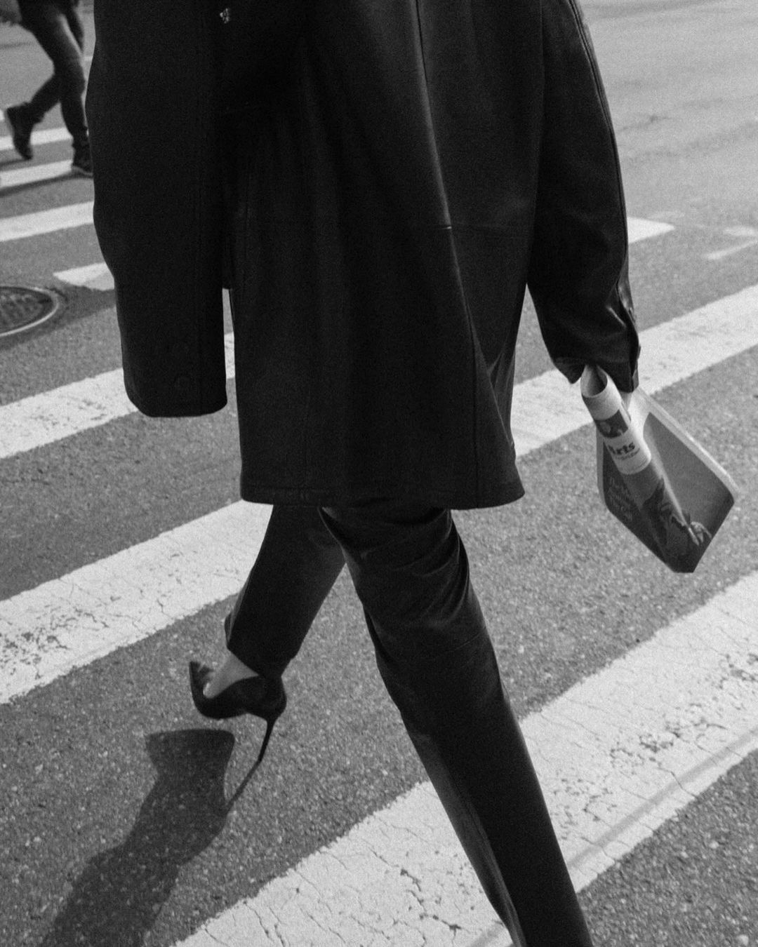 Monochrome image of a person in a dark coat and heels walking on a crosswalk, holding a newspaper, with other pedestrians in background.