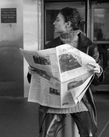 A woman in an overcoat and sweater is seen in an eye-level, medium shot as she reads a black and white newspaper indoors.