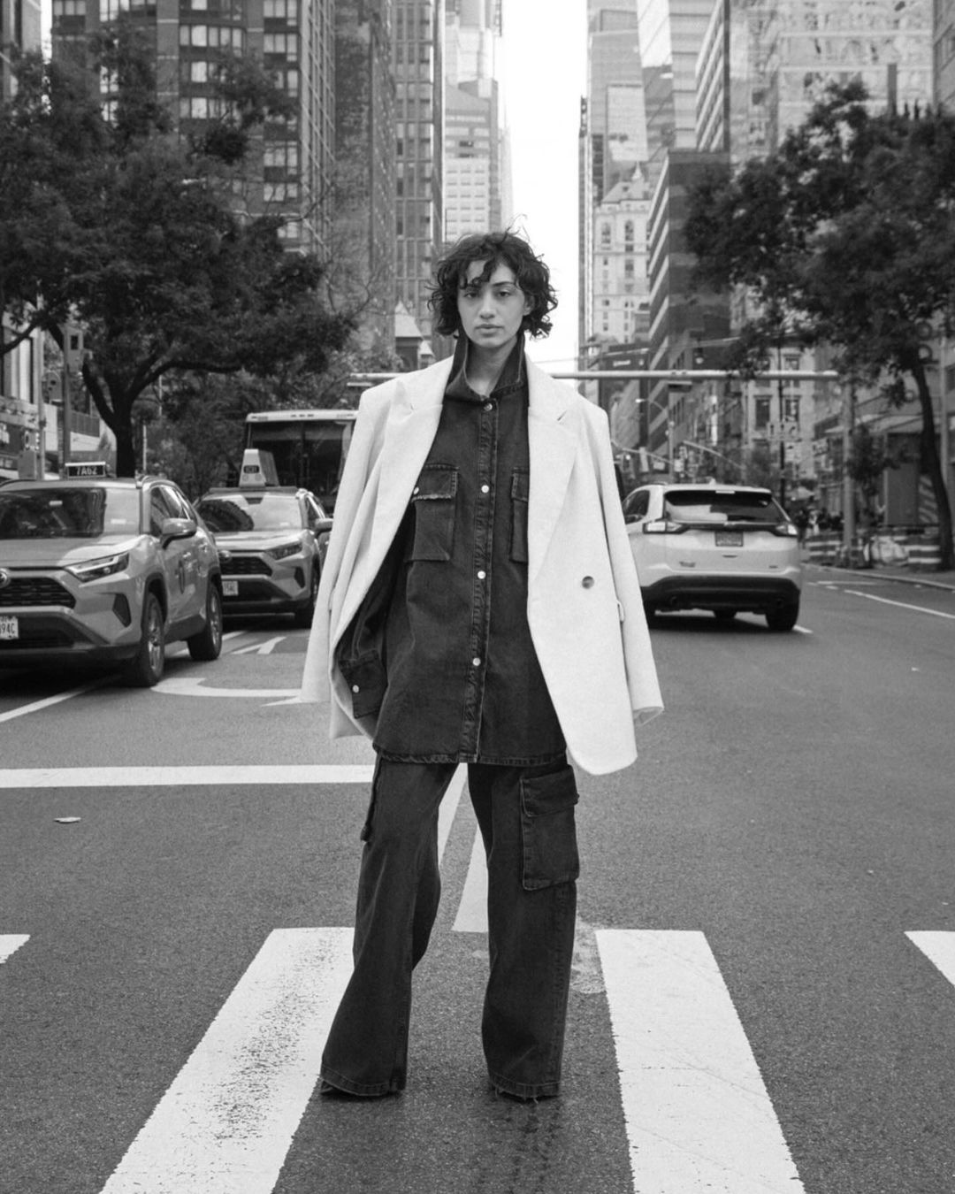 Woman in oversized denim and white blazer stands on a crosswalk in a black and white urban setting with cars and skyscrapers.