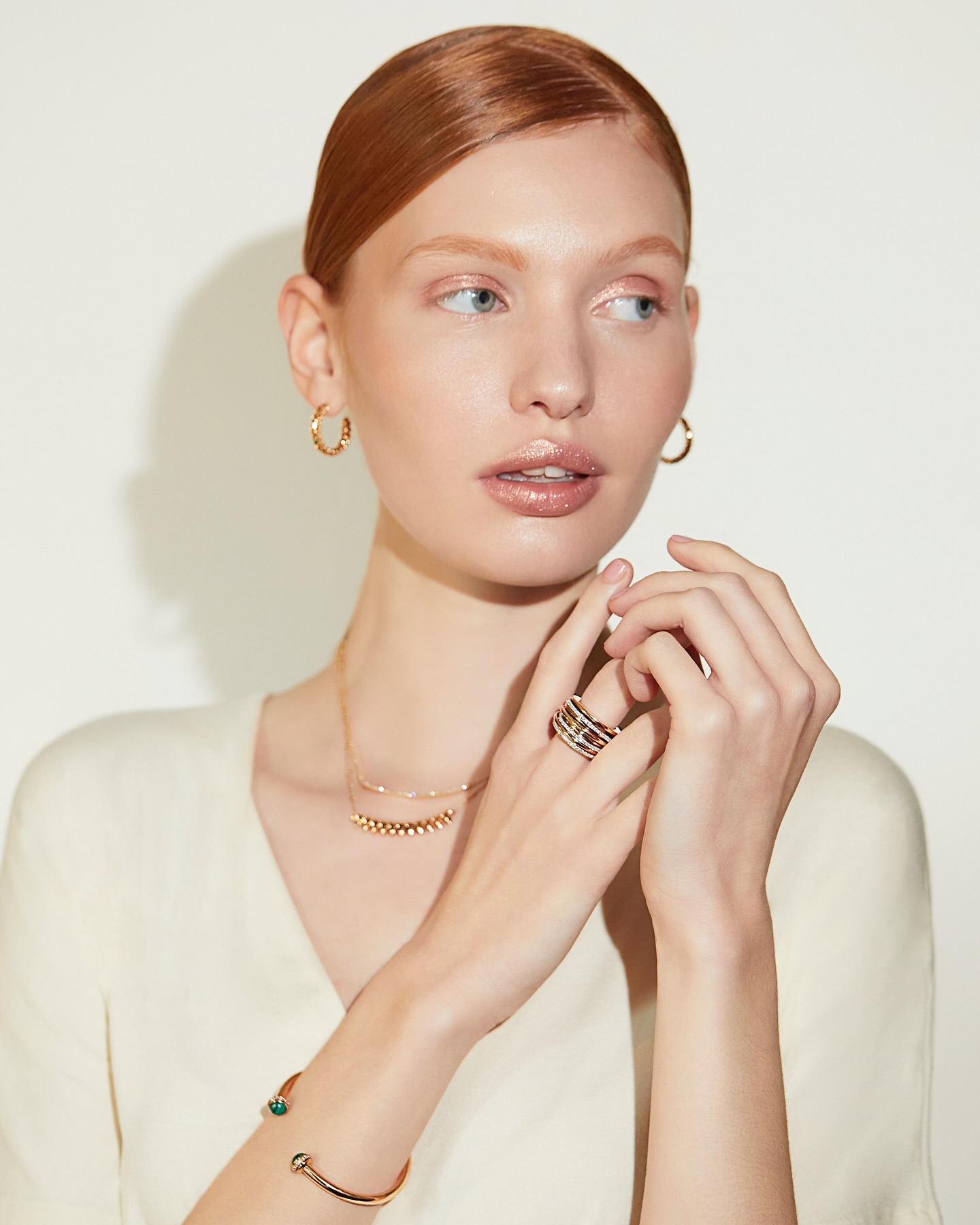 Red-haired woman wearing gold jewelry, hoop earrings, necklace, rings, and a bracelet against a neutral white background.