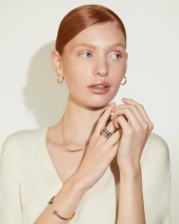 Red-haired woman wearing gold jewelry, hoop earrings, necklace, rings, and a bracelet against a neutral white background.