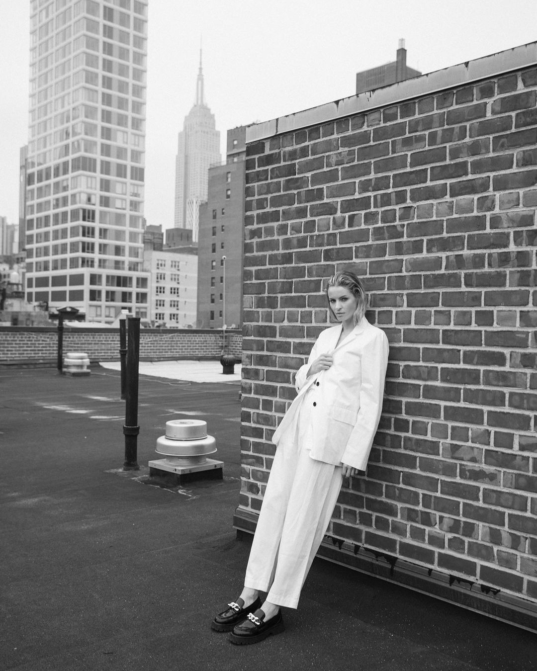A woman in a white suit leans against a brick wall on a rooftop in New York City with skyscrapers and the Empire State Building in the background.