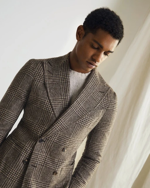 Man in a brown plaid double-breasted blazer and grey sweater looking down, posing against an off-white textured backdrop in a studio shot.