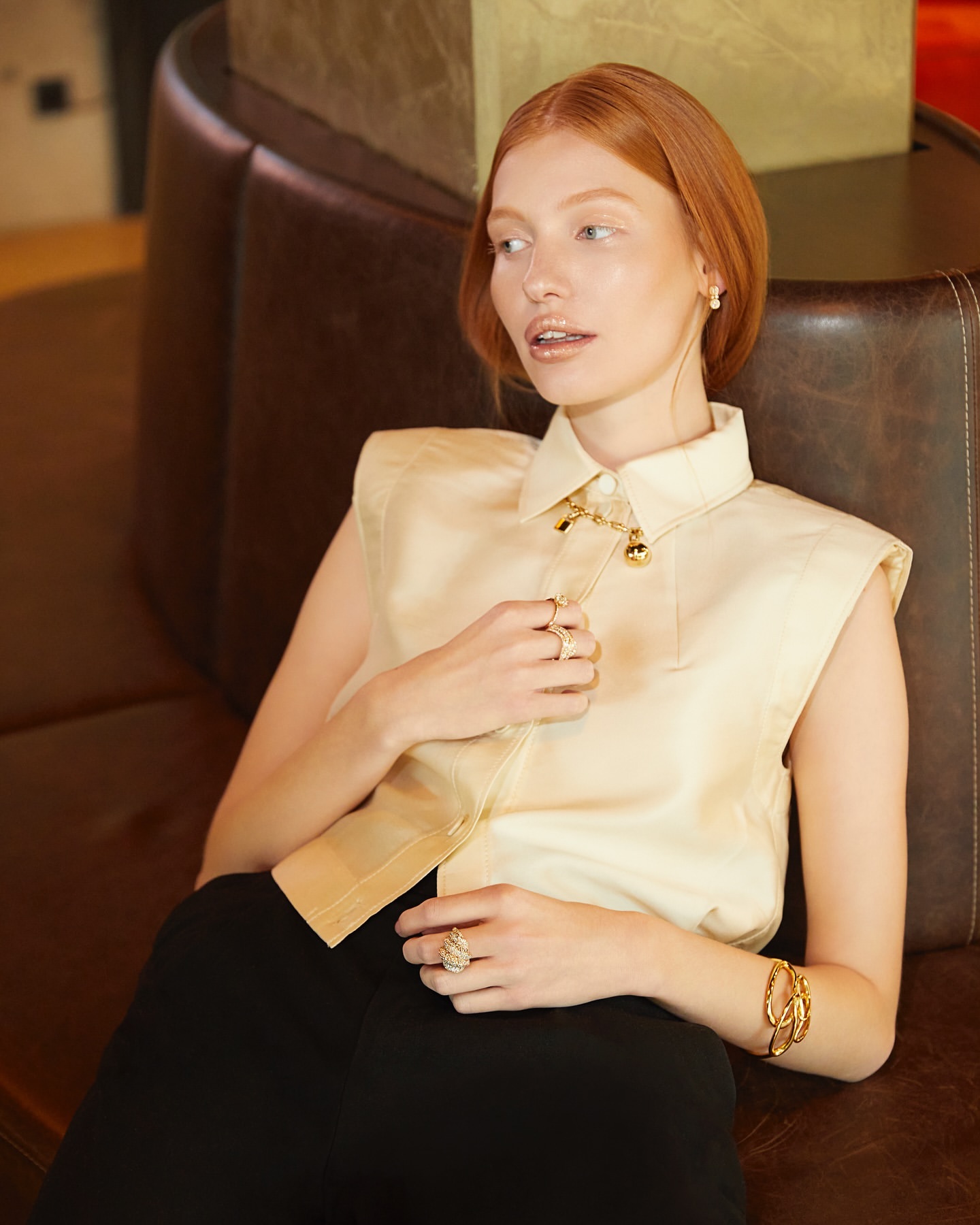 A red-haired woman wearing a sleeveless collared shirt and gold jewelry, sitting on a brown leather couch in an indoor setting.
