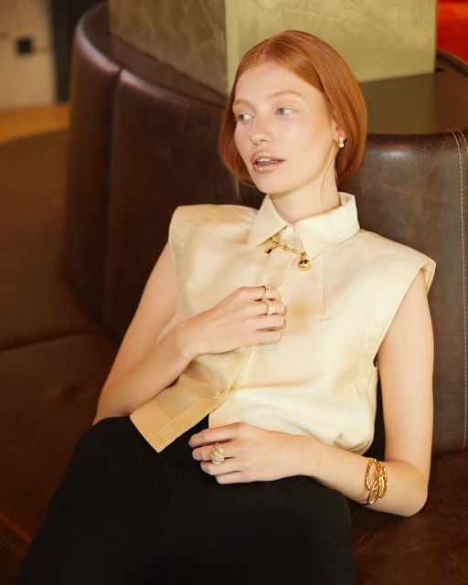 A red-haired woman wearing a sleeveless collared shirt and gold jewelry, sitting on a brown leather couch in an indoor setting.