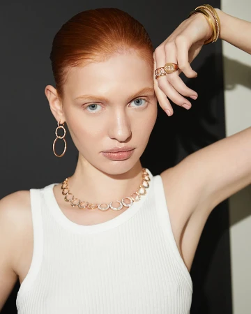 Red-haired model wearing gold jewelry including rings, necklaces, and hoop earrings with a white tank top.