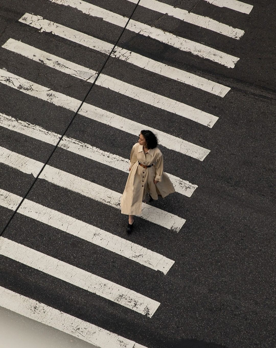 Aerial view of an adult female walking across a pedestrian crosswalk in a light beige trench coat and dark shoes on an asphalt street.