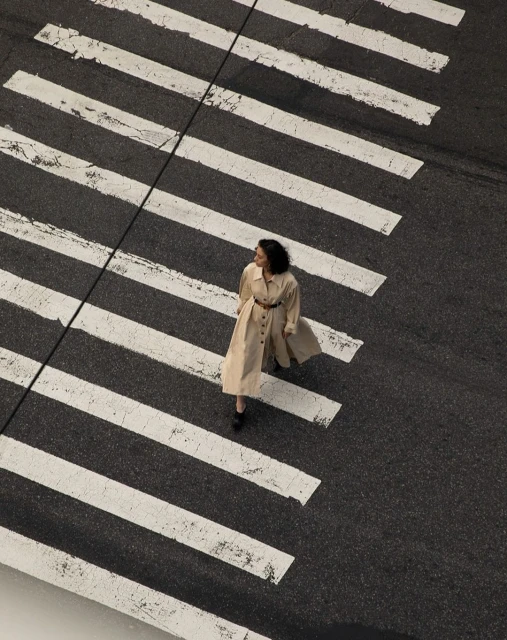 Aerial view of an adult female walking across a pedestrian crosswalk in a light beige trench coat and dark shoes on an asphalt street.