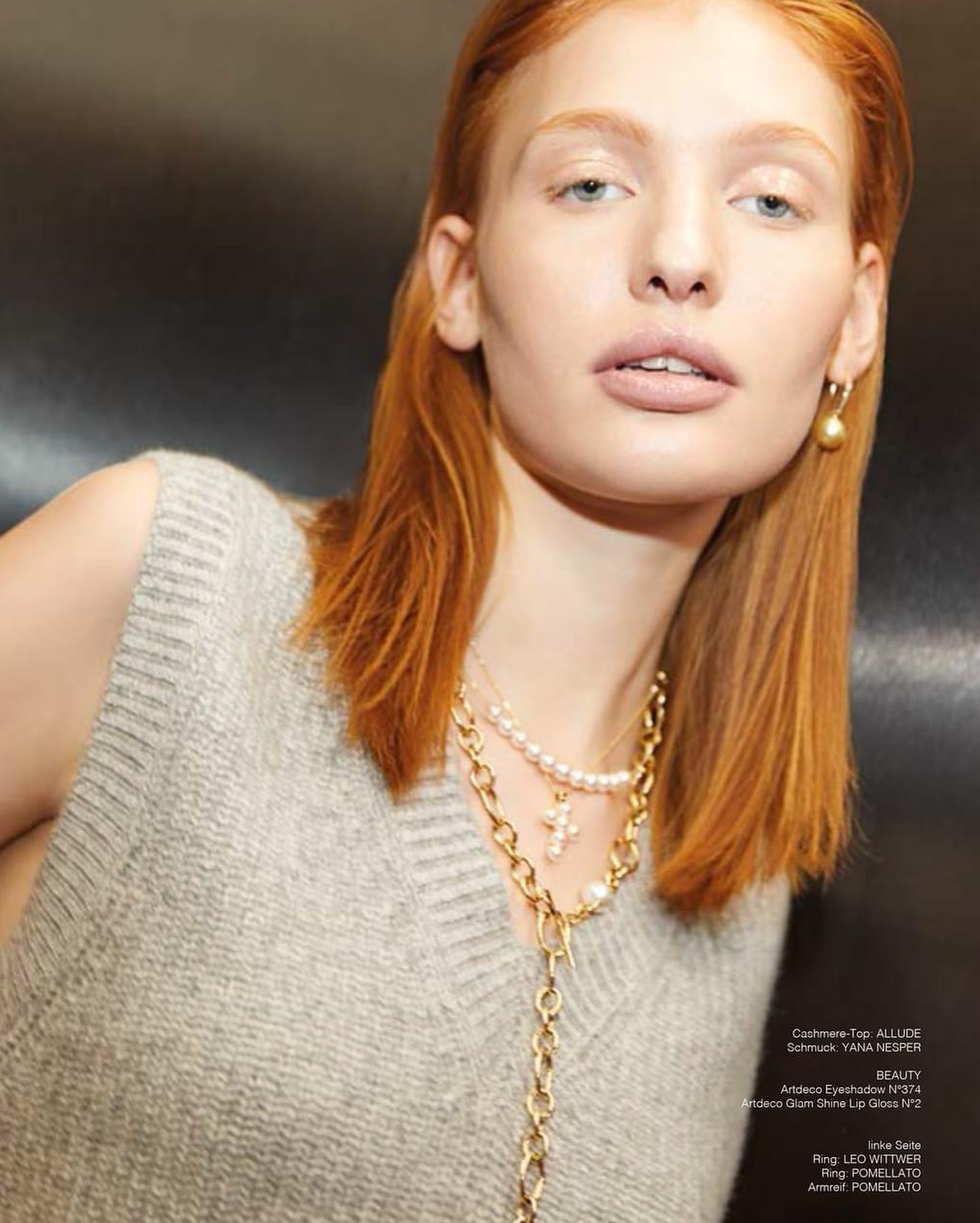Close-up of a red-haired woman in a gray sweater vest, accessorized with pearl and gold jewelry, against a dark background, indoor portrait.