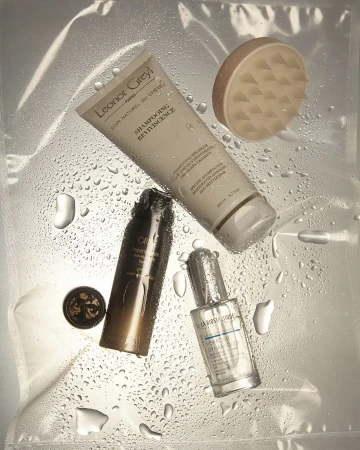 Still life of Leonor Greyl shampoo, Orbie hair product, and La Biosthetique styling oil on a clear, wet plastic surface.
