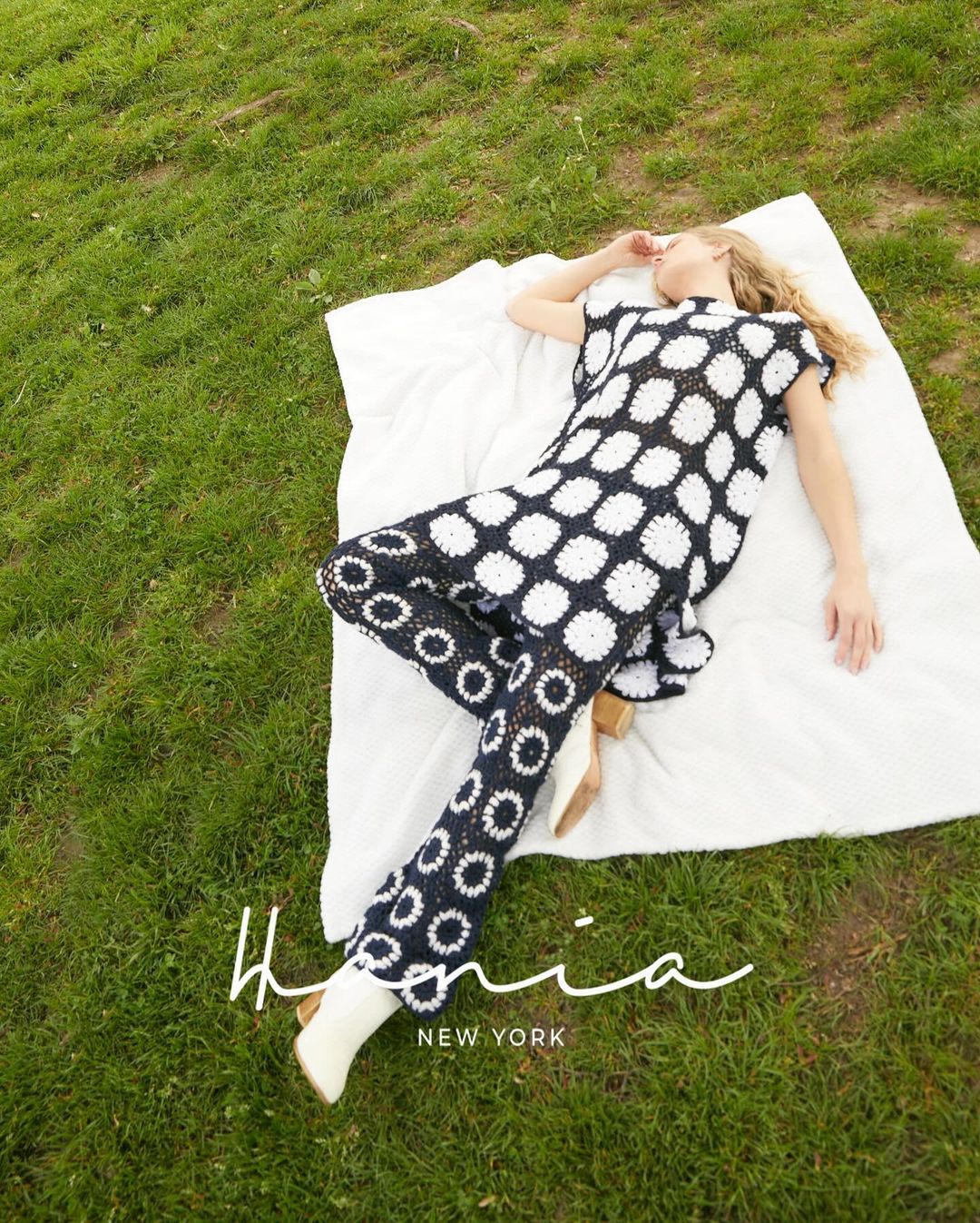 Woman in black and white patterned outfit lies on a white blanket on a green lawn, eyes closed in outdoor fashion shot.