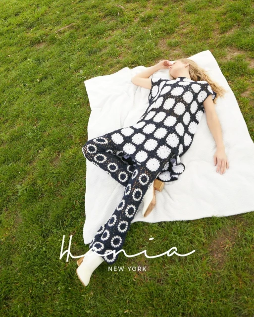 Woman in black and white patterned outfit lies on a white blanket on a green lawn, eyes closed in outdoor fashion shot.