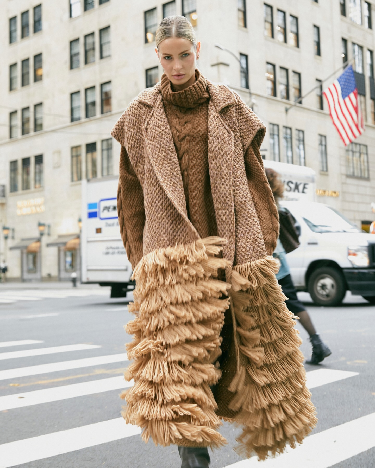 Fashionable adult female model wears a long brown knitted coat with fringe, posing on a crosswalk in a cityscape with vehicles.