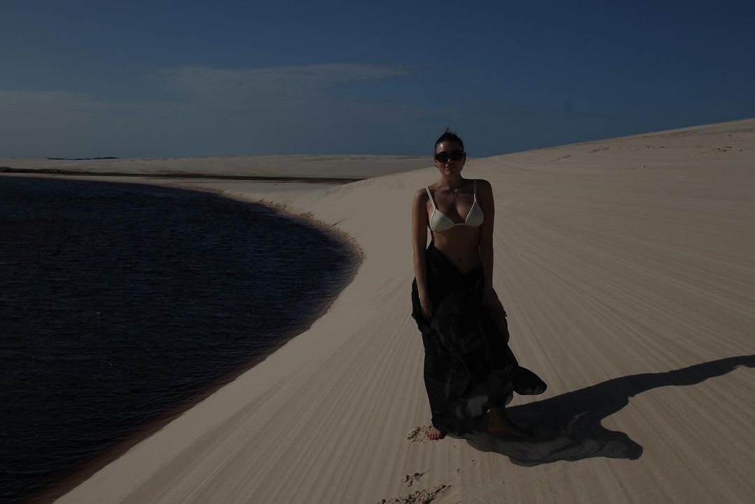 Woman in white bikini and black cover-up standing on a sand dune next to a pool of water under a clear blue sky in a desert landscape.