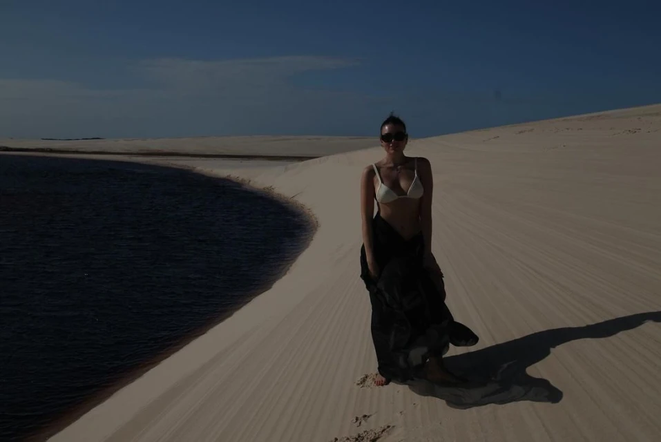 Woman in white bikini and black cover-up standing on a sand dune next to a pool of water under a clear blue sky in a desert landscape.