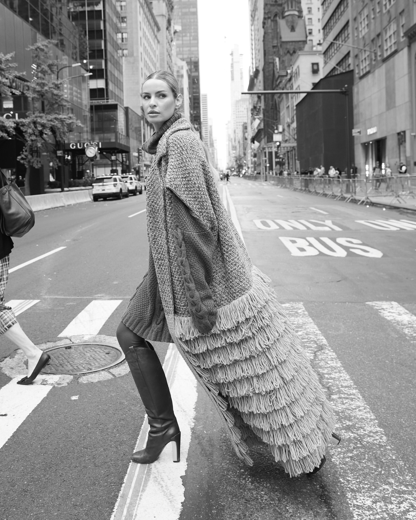 Adult woman poses in long sweater coat, crossing city street on crosswalk in black and white. Building and sky background.