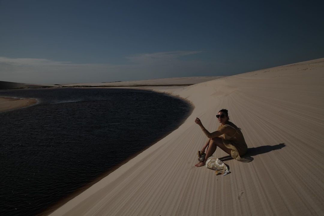 An adult woman wearing sunglasses and a dress sits on a sand dune next to a lake under a blue sky in an outdoor environment.