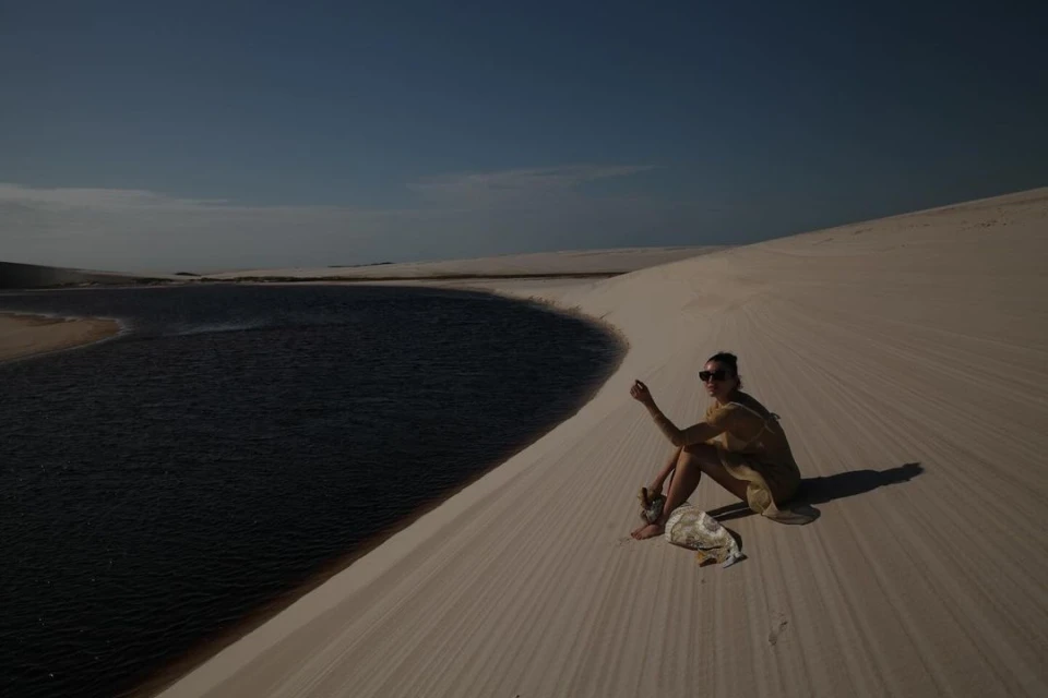 An adult woman wearing sunglasses and a dress sits on a sand dune next to a lake under a blue sky in an outdoor environment.