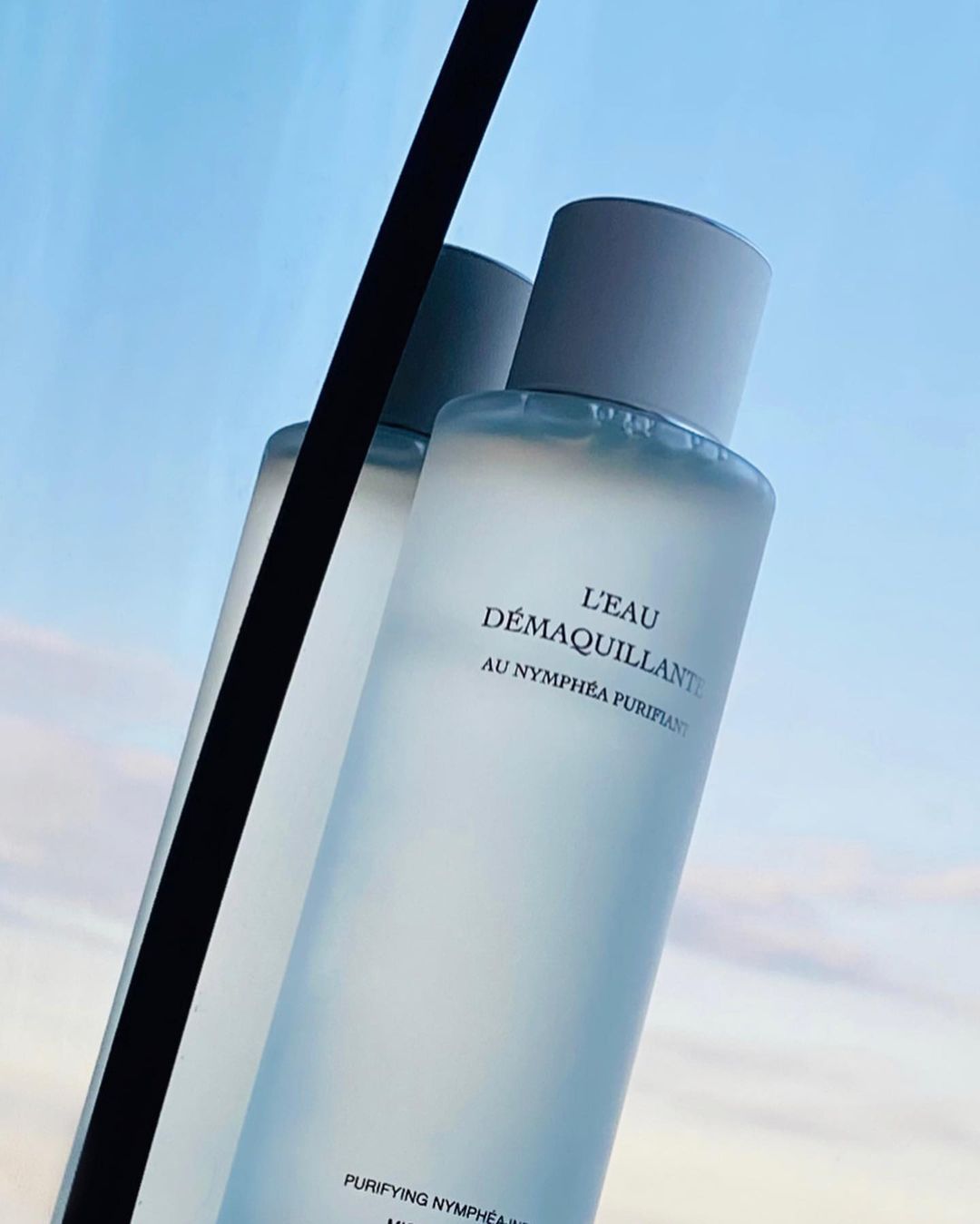 Bottle of L'Eau Demaquillante skincare product against a light blue sky, showcasing beauty and self-care in a minimalist composition.