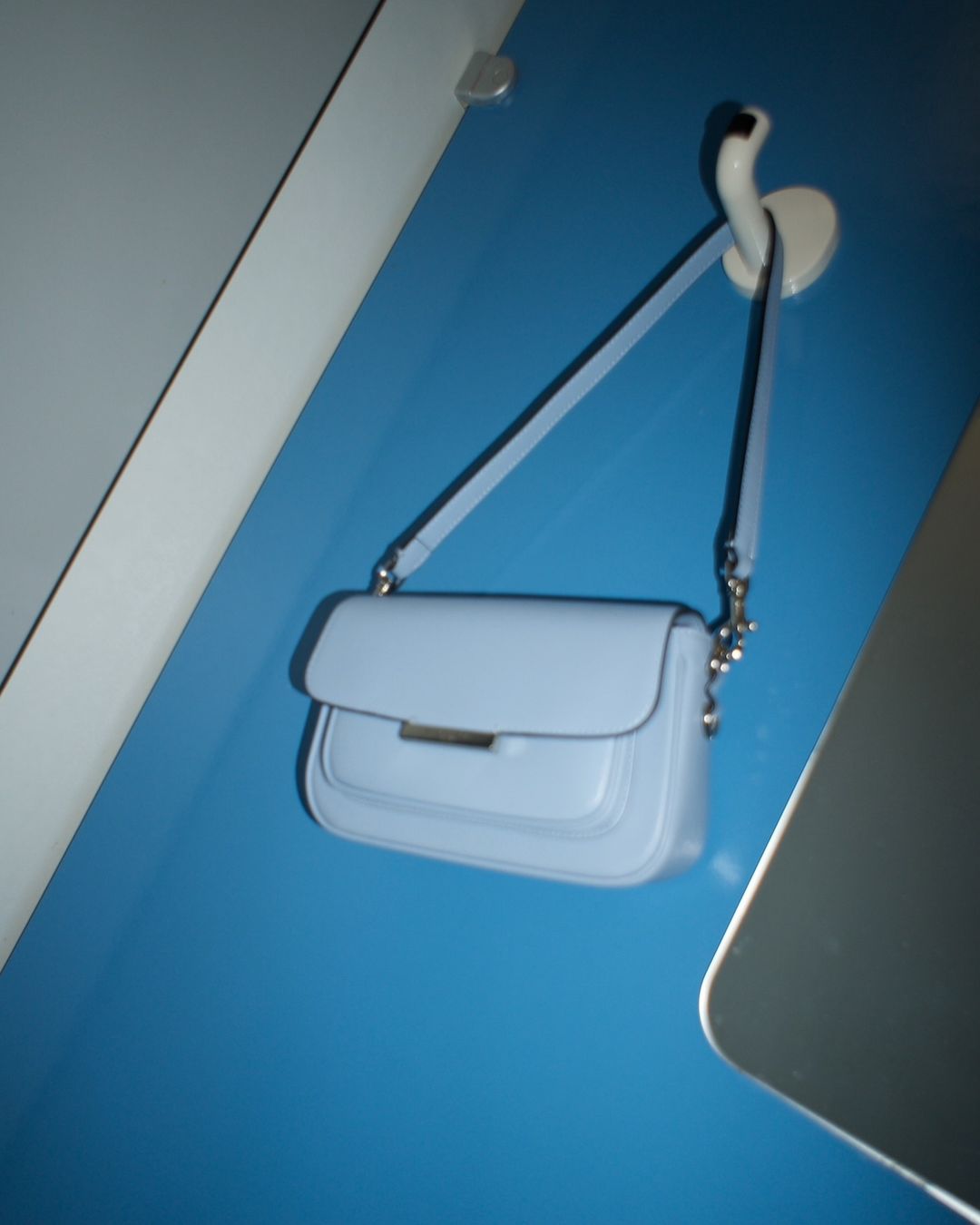 Light blue leather shoulder bag hanging from a white wall hook against a turquoise wall in an indoor setting, showcasing fashion.