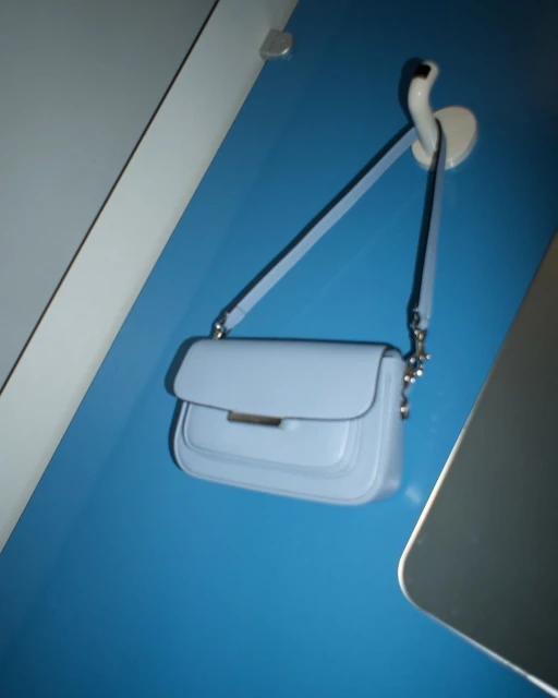 Light blue leather shoulder bag hanging from a white wall hook against a turquoise wall in an indoor setting, showcasing fashion.