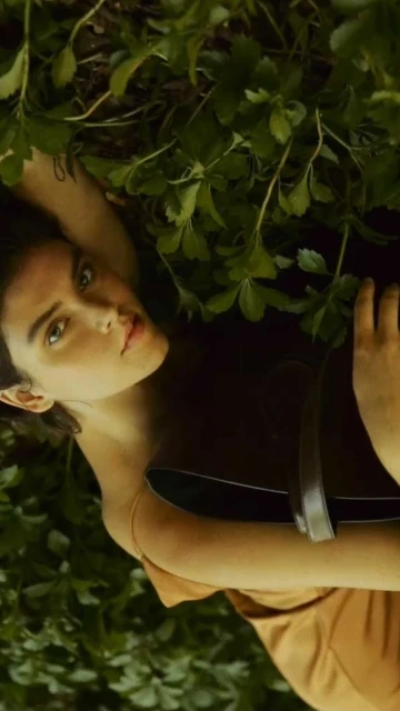 Brunette woman lies back among foliage wearing a tan dress and smiling up at the camera with fair skin and light-green eyes.