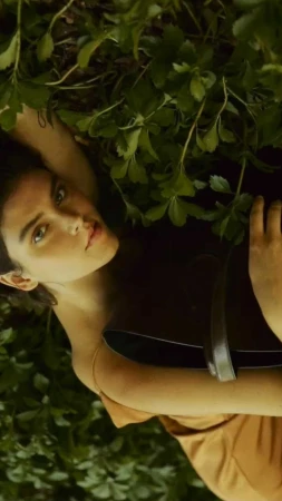 Brunette woman lies back among foliage wearing a tan dress and smiling up at the camera with fair skin and light-green eyes.