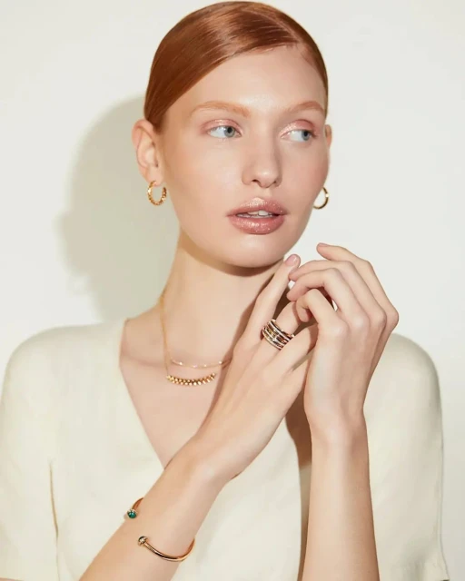 Elegant woman with auburn hair showcasing gold hoop earrings, a gold necklace, a bracelet, and a ring against a neutral backdrop.