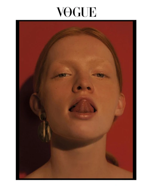 Close-up portrait of a fair-skinned young woman with an open mouth, tongue extended, wearing dangling earrings against a red background.