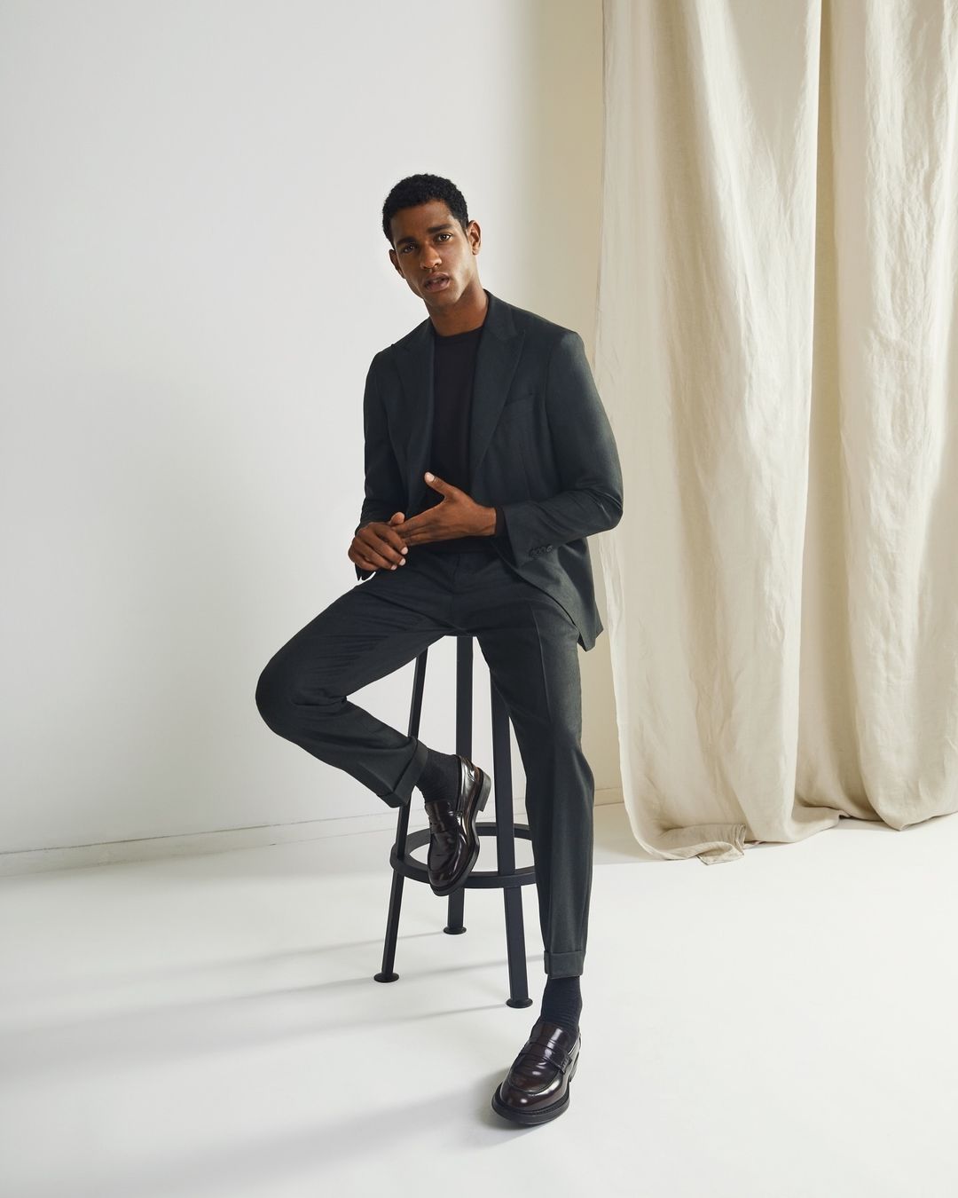 Young man in a dark tailored suit seated on a black metal stool against a white backdrop with cream curtains