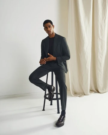 Young man in a dark tailored suit seated on a black metal stool against a white backdrop with cream curtains