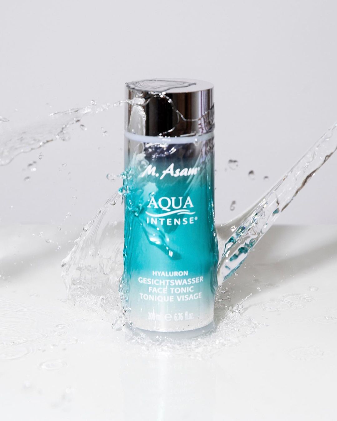 Product shot of M. Asam Aqua Intense face tonic bottle with water splashing around it, on a white background, studio lighting.