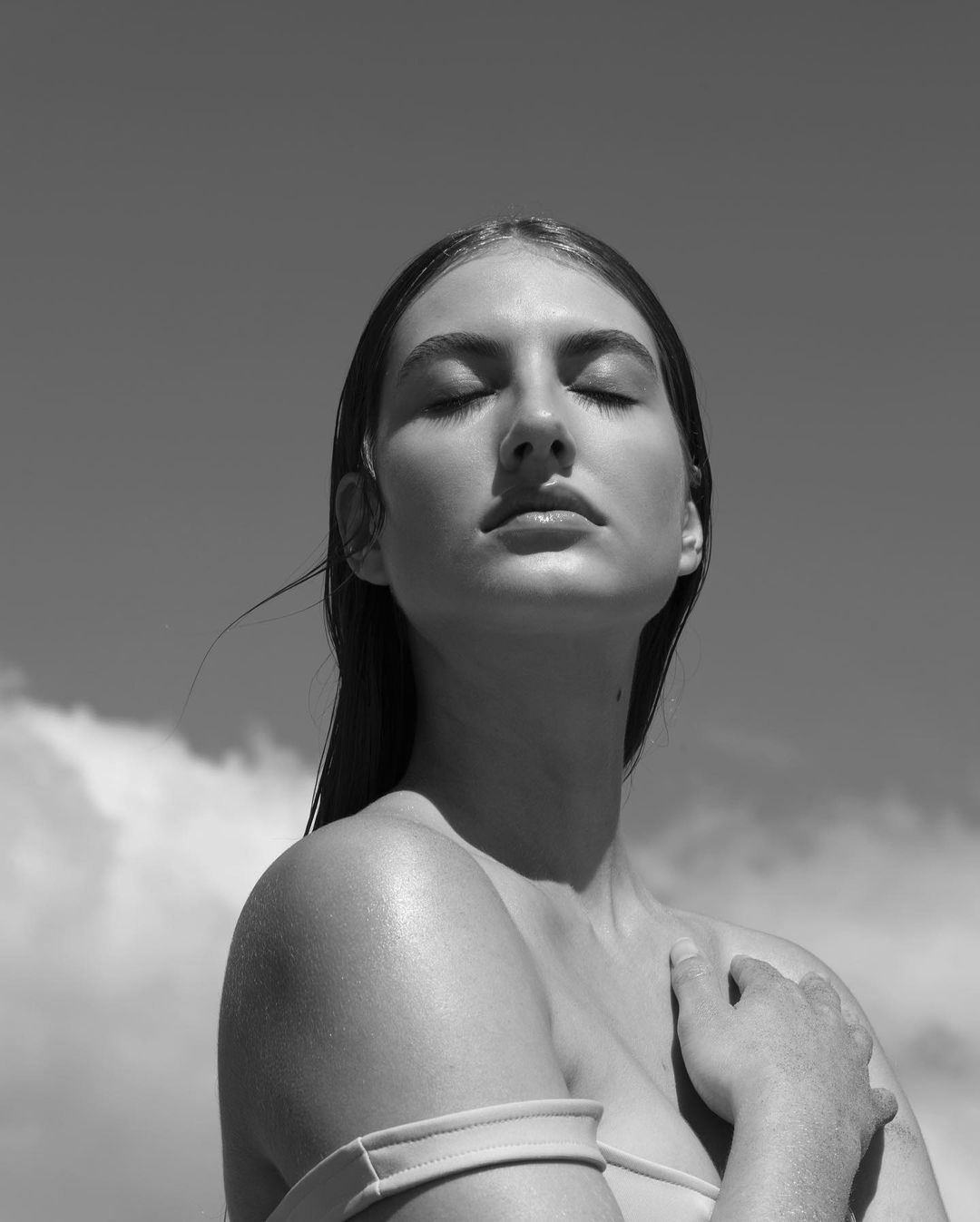 Black and white portrait of a young woman with closed eyes, wet hair, and bare shoulders against a cloudy sky in an outdoor setting.