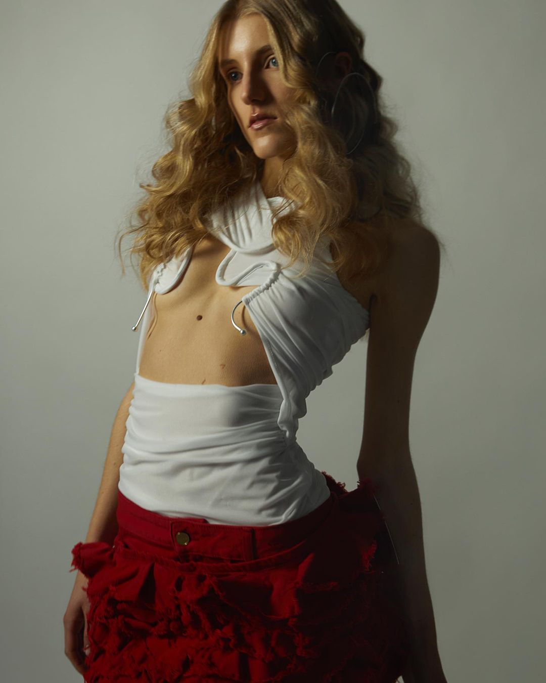 A fair-skinned woman with curly blonde hair poses in a white top with cutouts and a red, textured skirt against a light background.