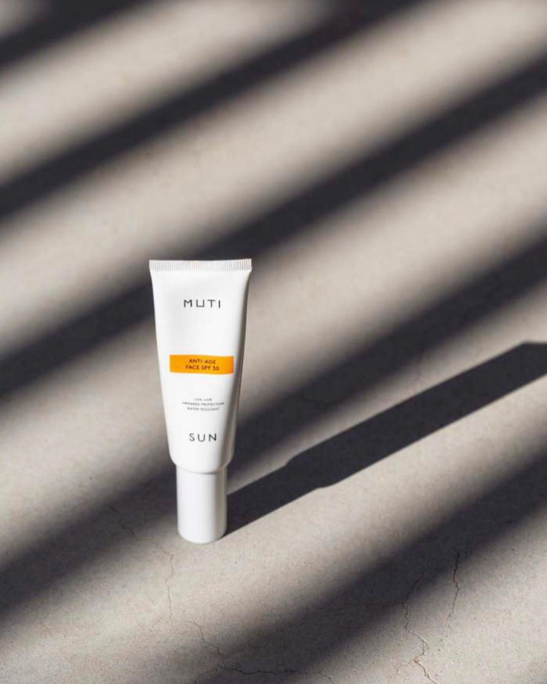 MUTI brand anti-aging face sunscreen SPF 50 tube product shot on a grey surface with shadow patterns from window blinds.